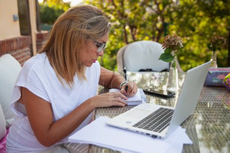Foto de Mujer concentrada teletrabajo con computadora y diario en el jardín de su casa - Imagen libre de derechos