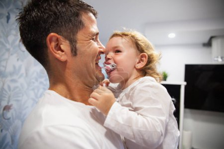 Photo for Happy father playing with his baby daughter's pacifier in the kitchen with laughter - Royalty Free Image