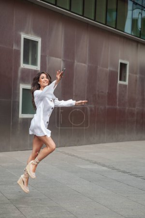 Photo for Young sporty girl jumping on the street happy with the mobile phone - Royalty Free Image