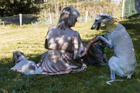 Foto de Mujer sentada sobre su espalda en el campo rodeada de sus mascotas, una es un galgo. - Imagen libre de derechos