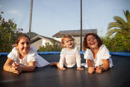 Téléchargez les photos : Trois petites filles soeurs allongées au soleil sur le trampoline souriant - en image libre de droit