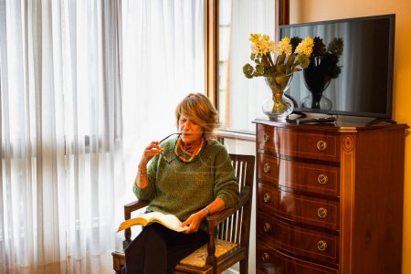 Photo for Mature woman sitting by window light enjoying reading - Royalty Free Image
