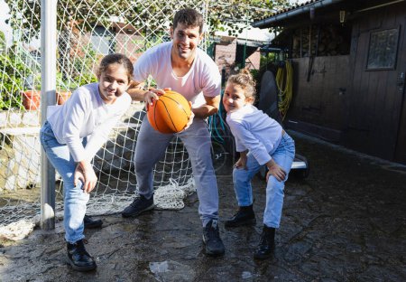 Téléchargez les photos : Jeune père souriant regardant la caméra avec ses deux filles et le basket-ball, à l'extérieur - en image libre de droit