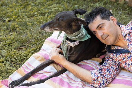 Téléchargez les photos : Portrait d'un homme détendu jouissant de son animal de compagnie couché sur le dos de son lévrier dans le jardin. - en image libre de droit