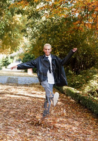 Téléchargez les photos : Jeune garçon moderne sautant entre les feuilles sur le sol dans le parc, photo verticale - en image libre de droit