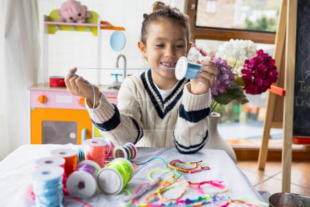 Foto de Retrato de una niña sonriente jugando manualidades con cordones de colores, haciendo pulseras. - Imagen libre de derechos