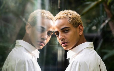 Photo for Portrait of beautiful young black boy with blond hair looking at camera reflected on glass of greenhouse in botanical garden - Royalty Free Image