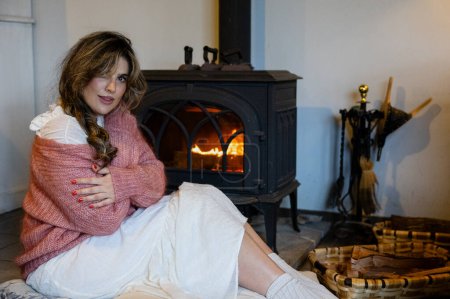 Téléchargez les photos : Portrait de femme assise devant la cheminée se détendre dans des vêtements confortables en regardant la caméra se détendre et se reposer - en image libre de droit