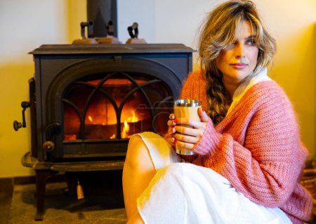 Foto de Retrato de una hermosa mujer sentada frente a la chimenea bebiendo una bebida caliente mientras se relaja - Imagen libre de derechos