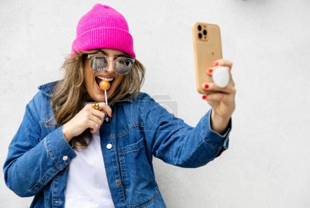 Foto de Retrato de chica sonriente con gafas y gorra disfrutando de una piruleta tomando una selfie, espacio en blanco - Imagen libre de derechos