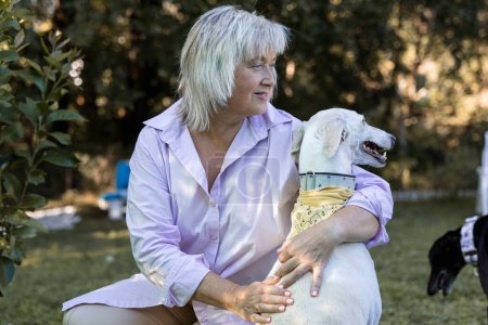 Téléchargez les photos : Relaxed mature woman hugging her white greyhound on a lazy day in the countryside. - en image libre de droit