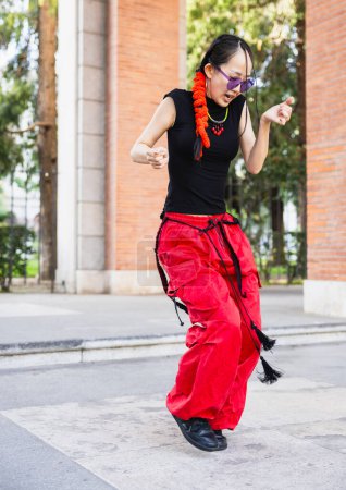Téléchargez les photos : "Un plan vibrant et dynamique capturant une jeune femme avec une tresse dansant dans la rue, portant un pantalon rouge baggy et des lunettes de soleil violettes à la mode." - en image libre de droit