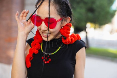 Téléchargez les photos : "Un cliché franc d'une danseuse de rue souriante tout en ajustant ses lunettes de soleil rouges en forme de cœur, ornées d'accessoires rouges vibrants. - en image libre de droit