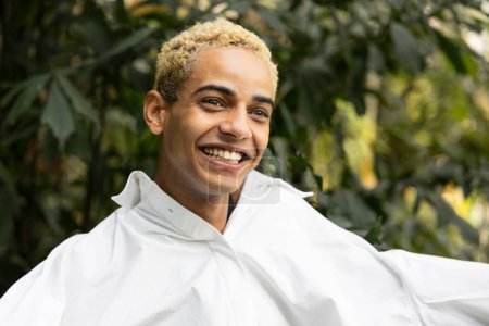 Foto de Fotografía franca de un joven feliz con el pelo rubio rizado, riéndose en una camisa blanca, ambientada en un fondo de denso follaje de jardín. - Imagen libre de derechos