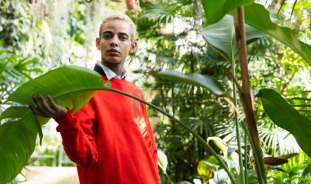 Téléchargez les photos : Un jeune homme aux cheveux blonds en pull rouge et chemise blanche pose dans un jardin botanique luxuriant et vert. - en image libre de droit