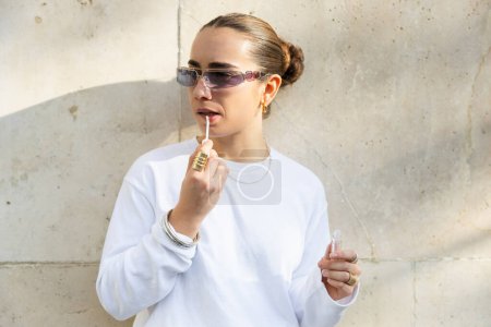 Foto de Una mujer de moda con una sudadera blanca aplica brillo labial mientras está de pie contra una pared envejecida, sus gafas de sol agregan a su comportamiento fresco. - Imagen libre de derechos