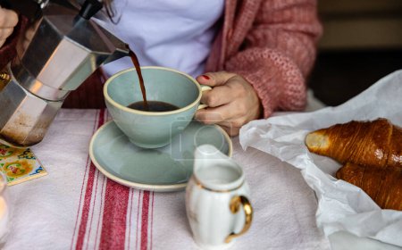 Foto de Primer plano de las manos vertiendo espresso caliente de una moka pot en una taza, con un delicioso croissant esperando. - Imagen libre de derechos
