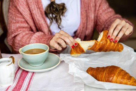 Foto de Una mujer en un suéter acogedor rasgando un croissant aparte con café en el lado, evocando un ambiente de desayuno relajado. - Imagen libre de derechos