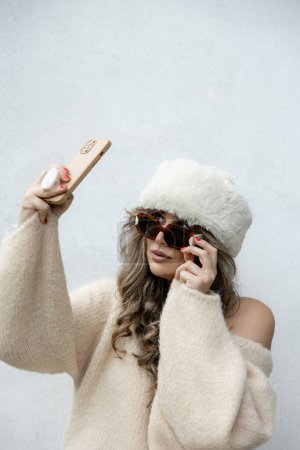 Téléchargez les photos : Une femme élégante dans un pull crème hors épaule et un chapeau de fourrure luxueux prend un selfie, exsudant élégance et charme d'hiver. - en image libre de droit