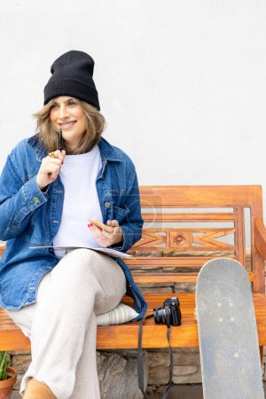Photo for Smiling artist enjoys a creative break outdoors, with a pen in hand and a smartphone for ideas, alongside her skateboard and camera. - Royalty Free Image