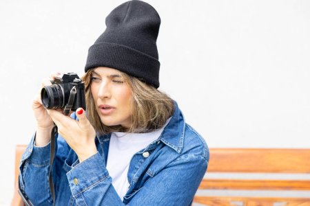 Photo for An engaged photographer with a vintage camera focuses intently on her subject, dressed in urban casual style with a denim jacket and beanie. - Royalty Free Image