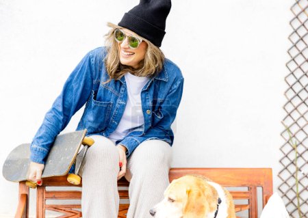 Foto de Una mujer alegre disfruta de un momento alegre en un banco con su monopatín, compartiendo una sonrisa con su fiel compañero beagle, exudando frescura urbana casual. - Imagen libre de derechos