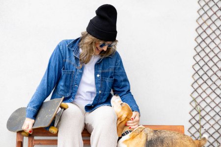 Foto de "Una joven alegre con una chaqueta de mezclilla y gorro disfruta de un momento lúdico con su perro beagle mientras está sentada en un banco al aire libre, sosteniendo un monopatín, mostrando un estilo de vida urbano relajado." - Imagen libre de derechos