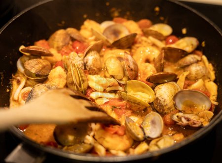 Foto de Un guiso de mariscos salados con almejas, camarones y tomates cocinando a fuego lento en una sartén negra. - Imagen libre de derechos