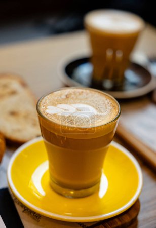 Foto de Un cálido café con leche artesanal con un hermoso arte de espuma, servido en una vibrante taza amarilla sobre una mesa de madera. - Imagen libre de derechos