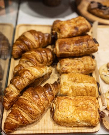 Foto de Croissants dorados y pasteles variados exhibidos en una bandeja de madera en una caja de panadería. - Imagen libre de derechos