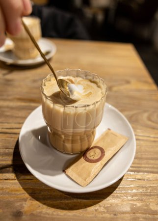 Foto de Disfrutando de un capuchino recién hecho, revolviendo la leche espumosa en una taza de vidrio transparente sobre una mesa de madera rústica. - Imagen libre de derechos