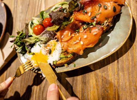 Téléchargez les photos : Une main coupée dans une assiette vibrante de pain grillé à l'avocat au saumon fumé avec un œuf parfaitement poché, accompagné d'une salade fraîche. - en image libre de droit