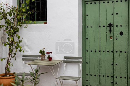Foto de "Una acogedora mesa al aire libre y sillas colocadas sobre el telón de fondo de una vibrante puerta verde y pared encalada en Frigiliana, España." - Imagen libre de derechos