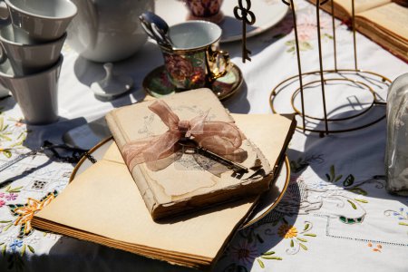 Foto de "Un primer plano de una fiesta de té vintage que muestra un libro antiguo atado con una cinta delicada, un par de gafas clásicas y una selección de tazas de té adornadas. - Imagen libre de derechos