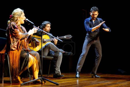 Foto de "Bailarín flamenco masculino intenso con gestos expresivos en la mano, acompañado por un cantante y guitarrista en una vibrante actuación en vivo." - Imagen libre de derechos