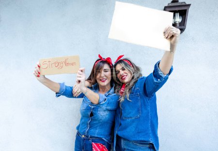 Zwei überschwängliche Frauen machen ein Selfie, eine hält ein Schild mit der Aufschrift "Starke Frauen" und feiert ihre Freude und Ermächtigung