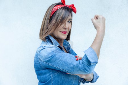 Photo for Modern day interpretation of the 'Rosie the Riveter' with a stylish woman showing her bicep in a denim outfit and red headscarf - Royalty Free Image