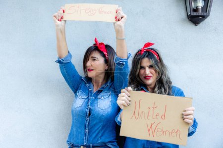 Zwei fröhliche Frauen in Jeans und rotem Kopftuch mit Schildern mit der Aufschrift "Starke Frauen" und "Vereinigte Frauen" vor grauem Hintergrund.