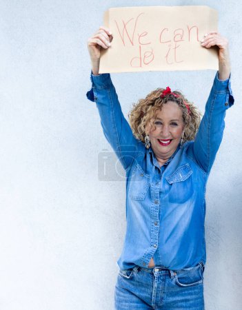 Photo for A smiling woman in blue denim holds up a 'We can do it' sign, embodying strength and positivity. - Royalty Free Image