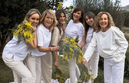 A happy group of women in white attire, holding bright yellow mimosa flowers, sharing a moment of camaraderie in nature.