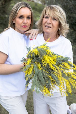 Portrait of two women sharing a heartfelt embrace, holding a vibrant bouquet of mimosa flowers, celebrating womanhood.