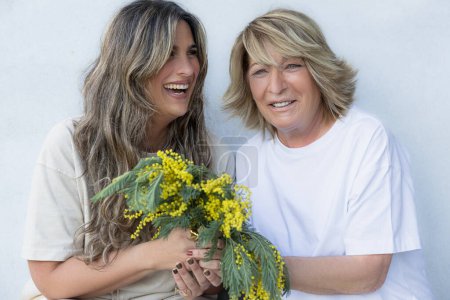 A spontaneous moment captures two women sharing laughter, with one holding a bouquet of mimosa flowers, embodying happiness and companionship.