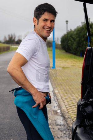 Téléchargez les photos : Jeune surfeur masculin sportif souriant à la caméra tout en se préparant à côté de sa voiture. - en image libre de droit