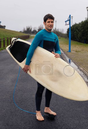 Surfer in a teal wetsuit carrying his board to the beach, with anticipation for the surf.