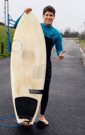 Téléchargez les photos : Surfeur souriant dans une combinaison bleue tient sa planche de surf, geste invitant vers la mer. - en image libre de droit