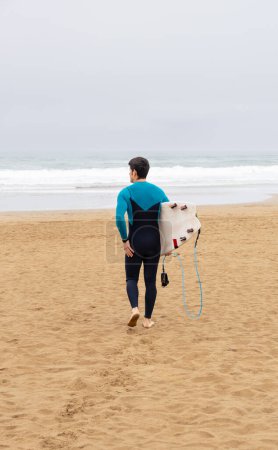 Rückansicht eines Surfers im blauen Neoprenanzug mit weißem Surfbrett, der an einem Sandstrand in Richtung Meer läuft.