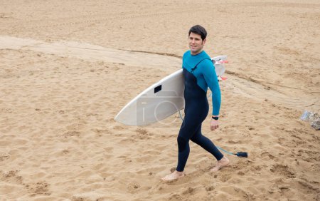 A young surfer in a blue and black wetsuit holding a surfboard, strolling on the sandy beach."
