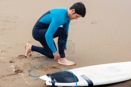 Téléchargez les photos : Jeune surfeur sur le rivage attachant sa planche de surf à sa cheville pour entrer dans la mer pour surfer - en image libre de droit