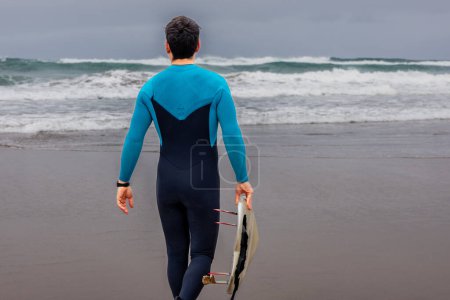Photo for Back view of a male surfer in a blue wetsuit carrying a surfboard, heading towards the sea under a cloudy sky. - Royalty Free Image