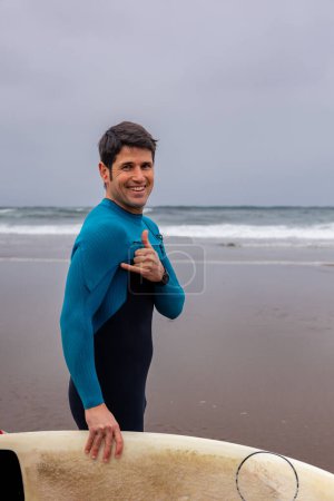 Ein gut gelaunter männlicher Surfer im blauen Neoprenanzug steht mit seinem Surfbrett und zeigt den Daumen nach oben an einem Sandstrand mit Wellen im Hintergrund..